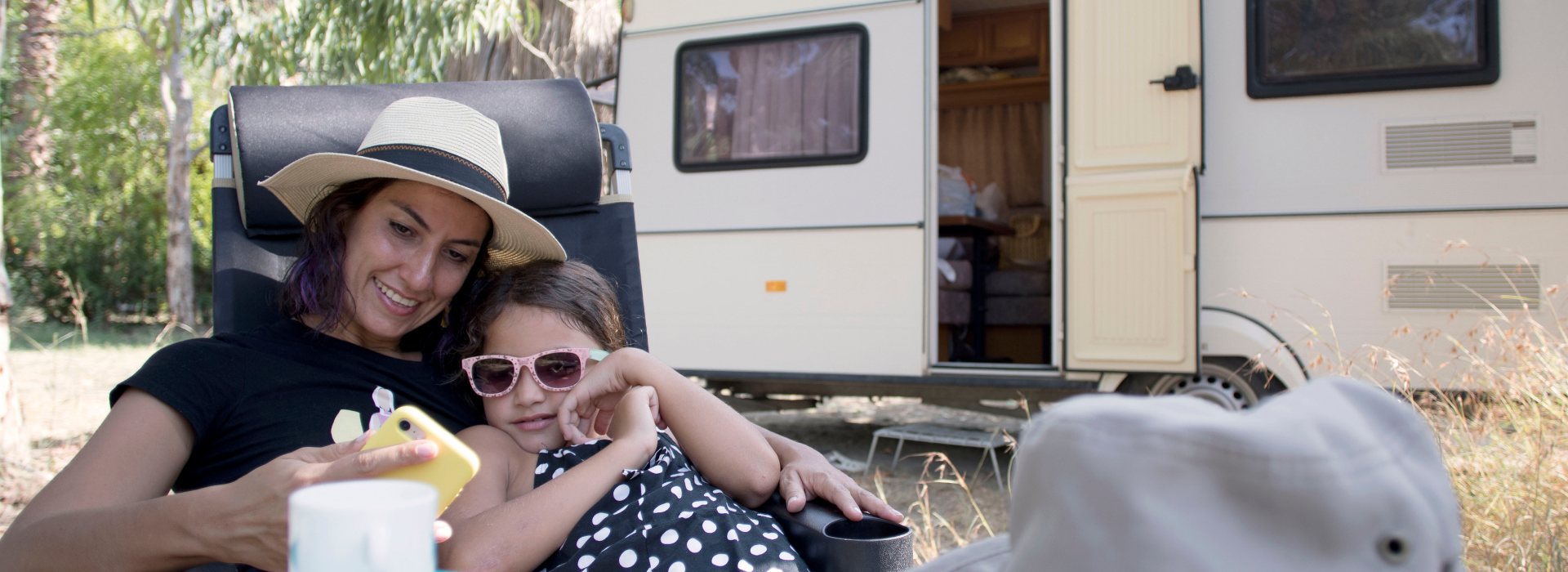 Woman and young girl lounge outdoors with camper parked behind them.