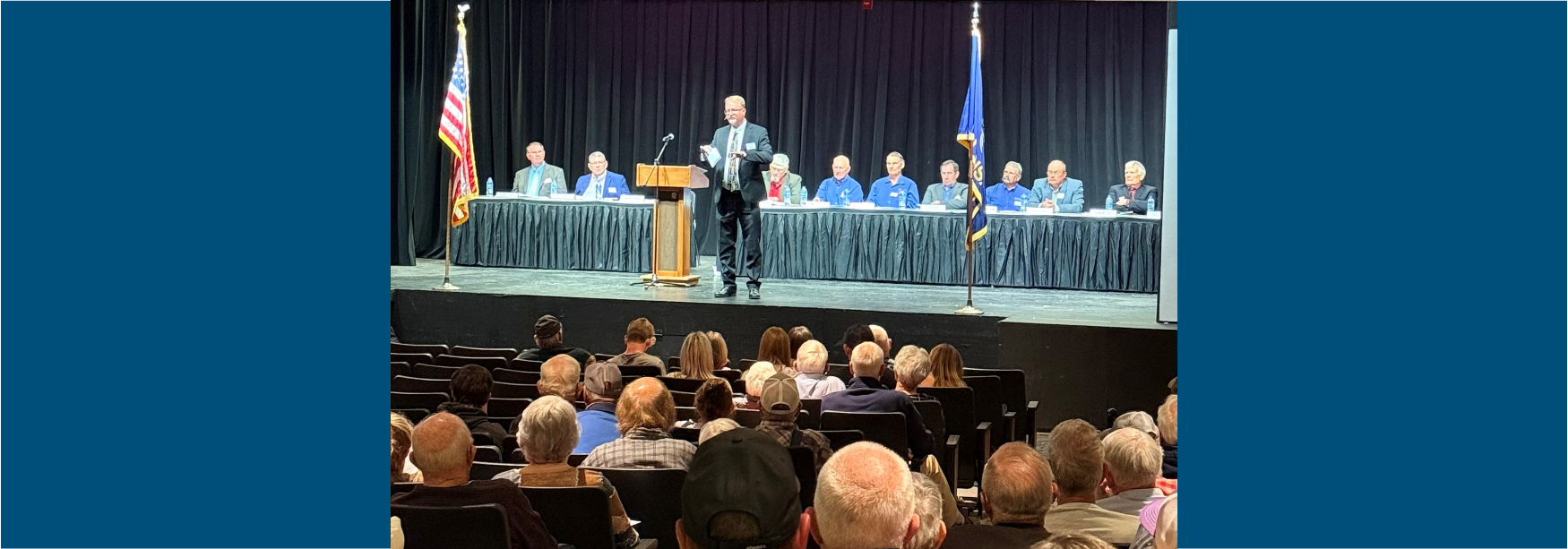 General manager on stage speaks to members in audience while board of trustees sits behind him.