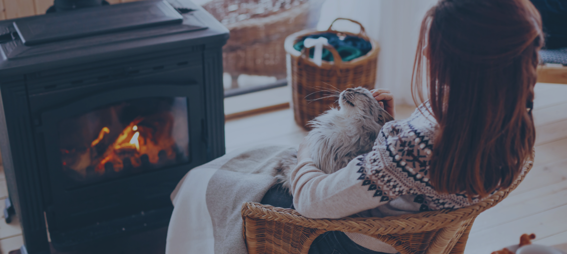 Brunette woman sitting in a chair with cat and blanket in her lap in front of a fireplace.